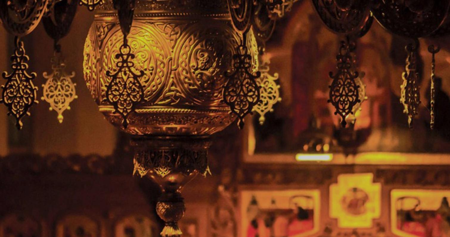 L&auml;hikuva koristeellisesta, kullatusta kattokruunusta, jossa on kauniita yksityiskohtia ja ornamentiikkaa. Taustalla n&auml;kyy himme&auml;sti valaistuna uskonnollinen maalauksia tai ikoneja kirkkomaisessa ymp&auml;rist&ouml;ss&auml;.